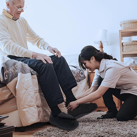 Older man seated on a bed while a caregiver helps adjust his black extra-wide comfort shoes.