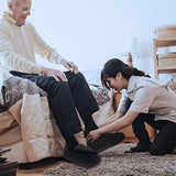 Older man seated on a bed while a caregiver helps adjust his black extra-wide comfort shoes.