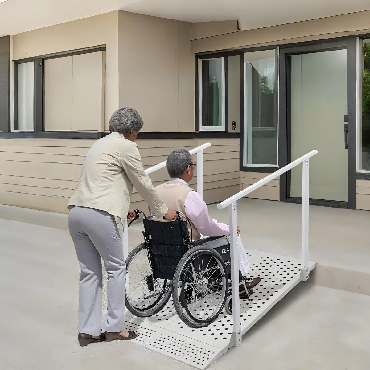 Caregiver pushing a wheelchair user up a white wheelchair ramp with handrails toward a building entrance.