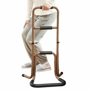 Person rising from a seated position while holding a brown portable stand assist with three foam-padded hand grips and a wide floor base.
