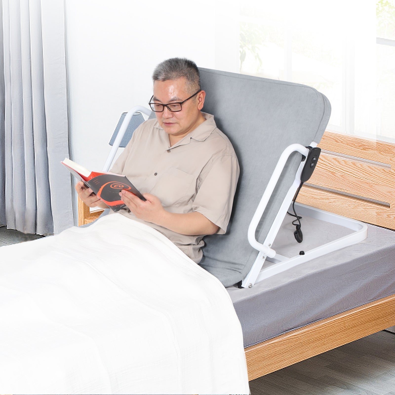 Man reading in bed while supported by a raised gray electric bed backrest, with side support rails and a wired controller visible.