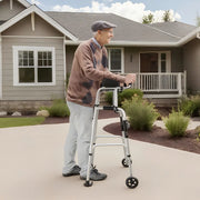 Older man using a silver upright walker with padded forearm supports on a sidewalk in front of a house.