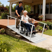 Man assisting an older woman in a wheelchair down an extended white ramp with handrails over the steps of a home porch.