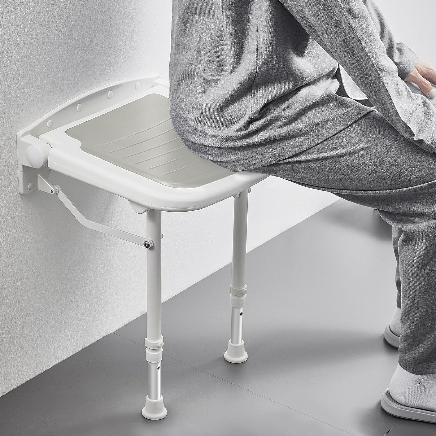 Person seated on a white wall-mounted folding shower seat in a tiled bathroom, showing the gray cushion, wall-mounted support frame, and adjustable legs.
