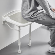 Person seated on a white wall-mounted folding shower seat in a tiled bathroom, showing the gray cushion, wall-mounted support frame, and adjustable legs.
