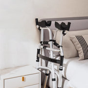 Silver upright walker folded for storage beside a bed, showing its compact frame, padded forearm supports, and hand grips.