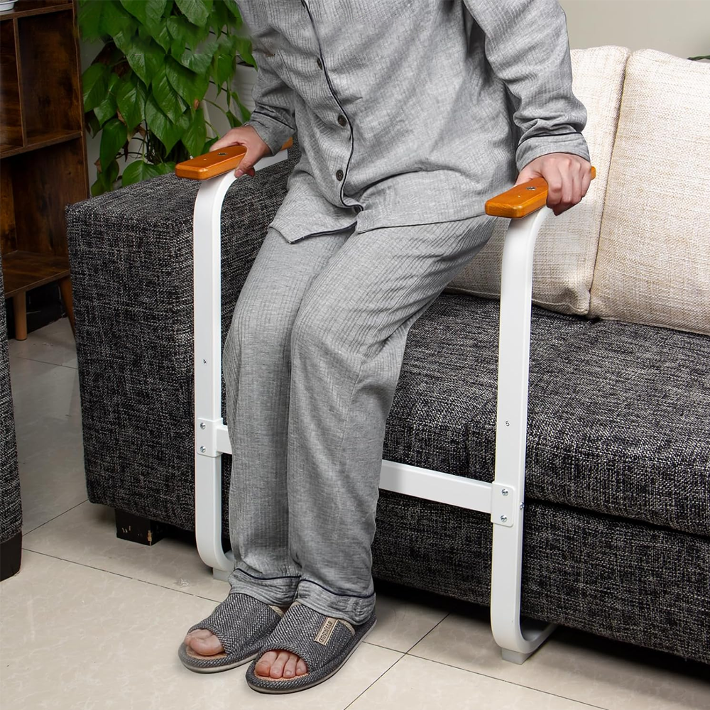 Person seated on the edge of a sofa gripping the wooden handles of a sit-to-stand assist frame, with the tray removed for standing support.