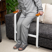 Person seated on the edge of a sofa gripping the wooden handles of a sit-to-stand assist frame, with the tray removed for standing support.